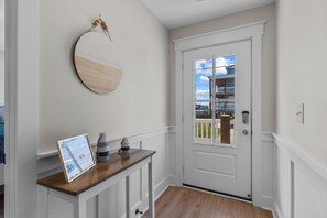 A hallway with a white door, a wooden console table with a framed picture, and a round wall hanging. - Field of Dreams #177 - 3BD Beach Cottage, Steps from the Ocean! (North Topsail Beach)