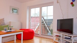 A modern living room with a large window, a red bean bag chair, a wooden table, a TV on a white stand, and a bookshelf. - Apartment Raum in Fehmarn Orth (Fehmarn)
