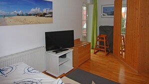 A room with a bed, a TV on a white stand, a wooden cabinet, a green chair, and a wooden door. - Apartment Raum in Fehmarn Orth (Fehmarn)