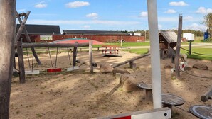 A playground with swings, a slide, and a red-roofed building in the background. - Stunning Cabin in Camping Resort (Lokken)