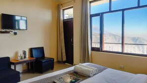 Una habitación de hotel con una cama, un televisor montado en la pared, una silla, una mesita con una bandeja y una ventana con vistas a la montaña.