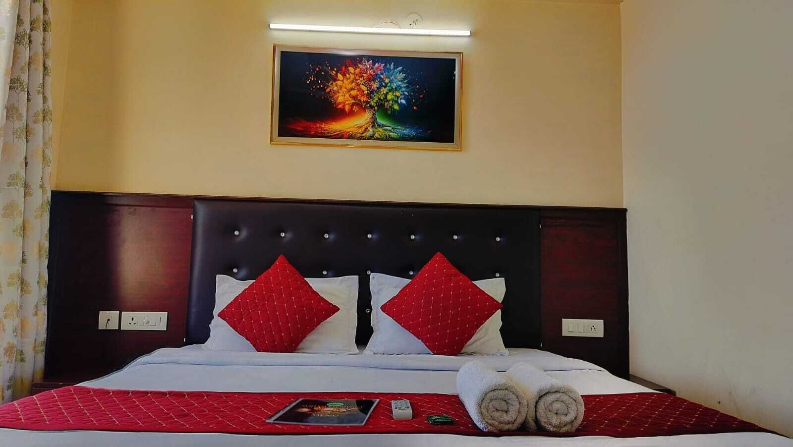 Habitación de hotel con una cama, ropa de cama roja y blanca, dos almohadas rojas, un cuadro en la pared y una mesita de noche con un control remoto y un libro.