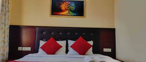 Habitación de hotel con una cama, ropa de cama roja y blanca, dos almohadas rojas, un cuadro en la pared y una mesita de noche con un control remoto y un libro.