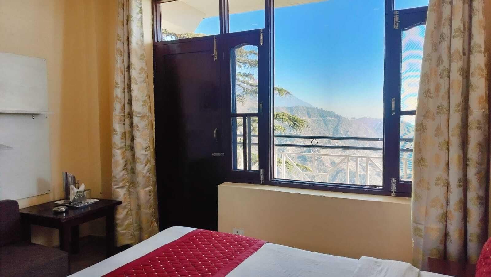 Habitación de hotel con cama, una mesita de noche con lámpara, ventana con vistas a montañas y cortinas.
