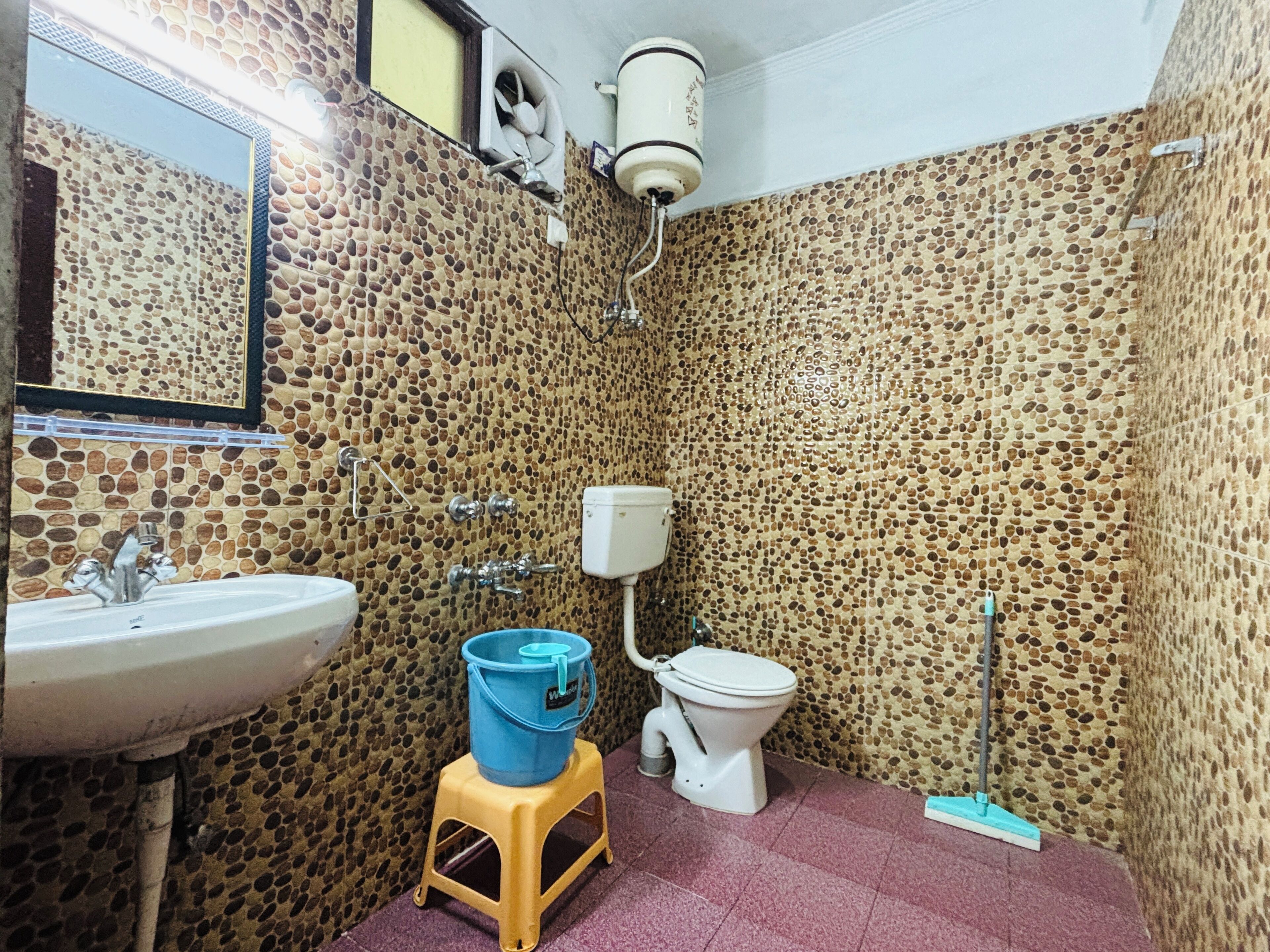 A bathroom with a stone-patterned wall, a white sink, a toilet, and a blue bucket on a stool.