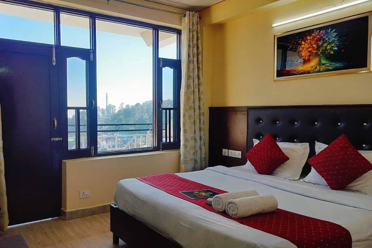A hotel room with a large bed, red pillows, a red bedspread, a painting on the wall, and a view of a balcony and cityscape through a large window.