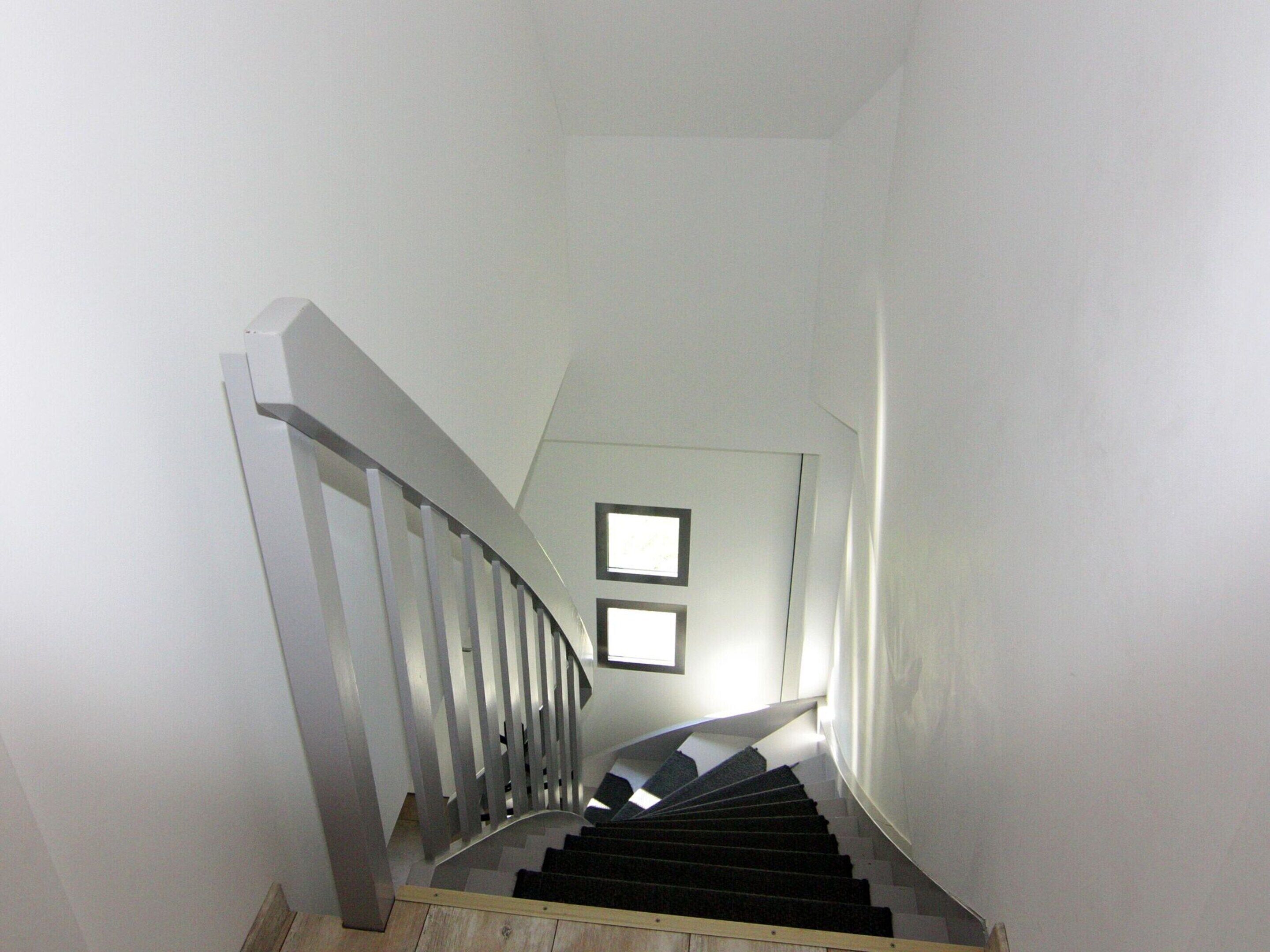 Un escalier avec une rampe en bois et des marches noires, menant à un palier où sont accrochées deux images encadrées au mur.