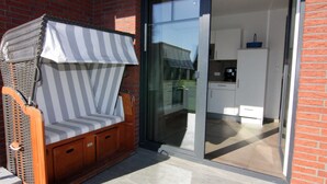 Een rieten stoel met een gestreept kussen op een terras met een glazen deur naar een keuken.