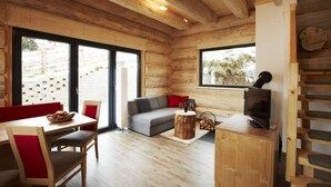 Interior acogedor de una cabaña con techo de madera, un sofá, una mesa de comedor con sillas, un televisor y un ventanal con vistas al exterior.