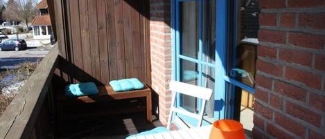 A wooden deck with a bench and a table, a blue chair, and a brick wall with a window.