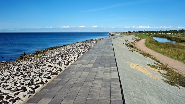 Un chemin pavé longe une zone côtière caractérisée par une rive rocheuse et une étendue d’eau.