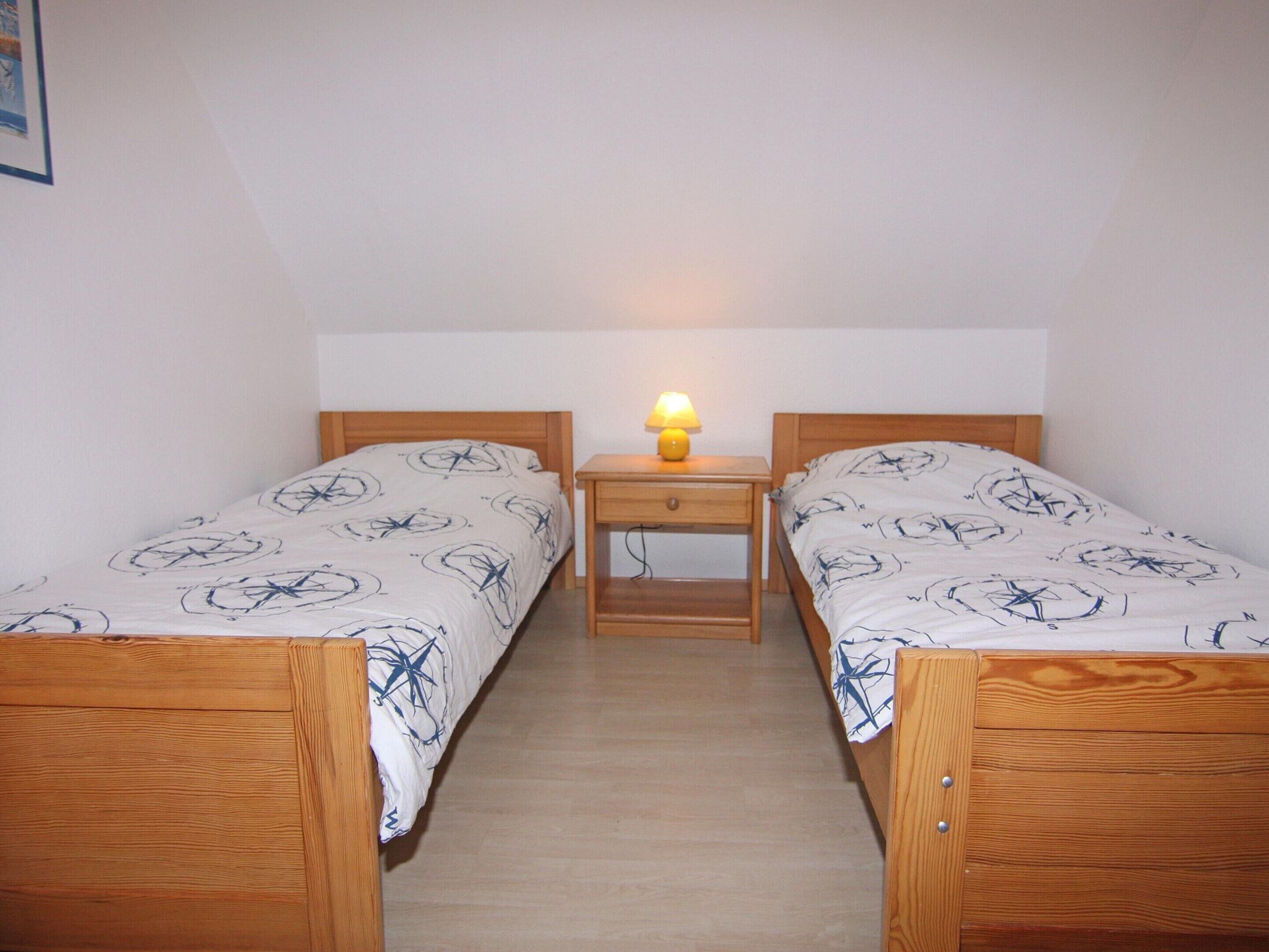 Une petite pièce simple, avec deux lits en bois, une table de chevet avec une lampe et un mur blanc uni.