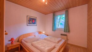 Een slaapkamer met een houten plafond, een bed met twee kussens, een nachtkastje met een lamp en een raam met gordijnen.