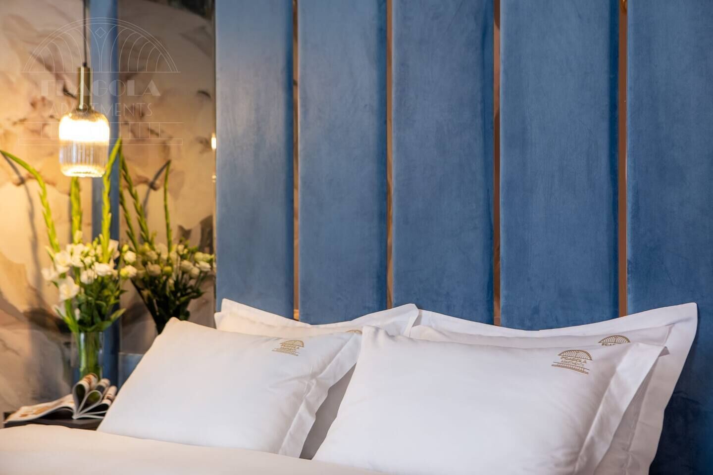 A bed with white linens and a blue headboard, a hanging light fixture, and a vase with flowers.
