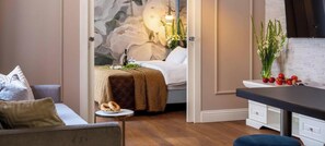 A modern hotel room with a bed, a small round table with snacks, a kitchen area, and a large floral painting on the wall.