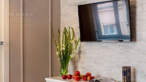 Una habitació amb un televisor fixat a una paret de maó, un vas amb flors, pomes i llibres damunt d'una taula blanca.