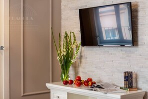 A room with a TV mounted on a brick wall, a vase with flowers, apples, and books on a white table.