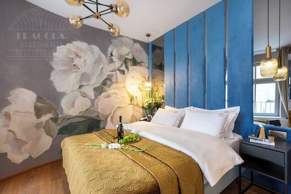 Un dormitorio con un gran mural floral, una cama con colcha dorada, una mesita de noche con una botella de vino y copas, y una pared azul decorativa.