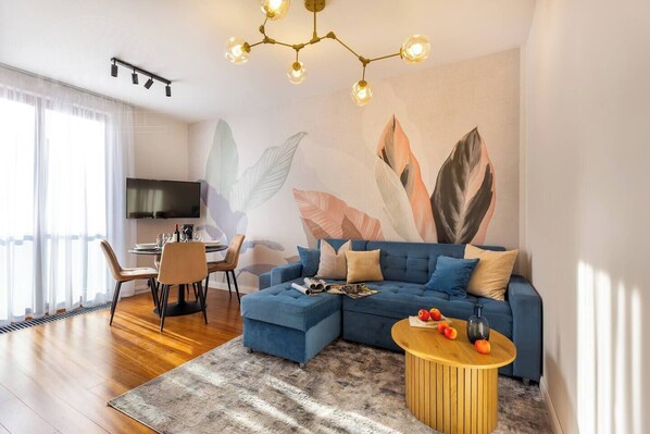 Un salon moderne comprenant un canapé d’angle, une table à manger avec des chaises, un téléviseur à écran plat fixé au mur et une grande fresque murale abstraite représentant des feuilles.