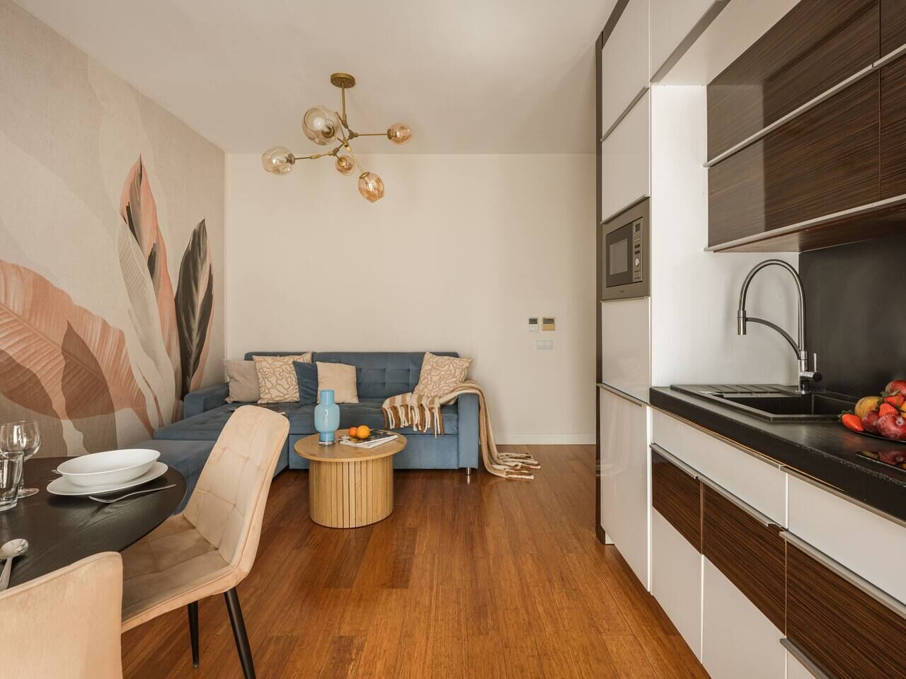 A modern kitchen with a built-in oven, a dining area with a round table and chairs, and a living area with a sofa and a decorative wall.