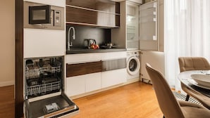Cocina compacta con lavavajillas, microondas y lavadora, zona de comedor con mesa y sillas, y ventana con cortinas traslúcidas.