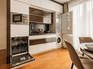 A compact kitchen with a dishwasher, microwave, and washing machine, a dining area with a table and chairs, and a window with sheer curtains.