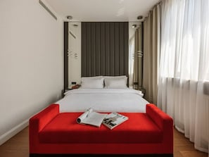 Ein Hotelzimmer mit einem Bett, einer roten Sitzbank, einer Zeitschrift, einem Spiegel und Vorhängen.