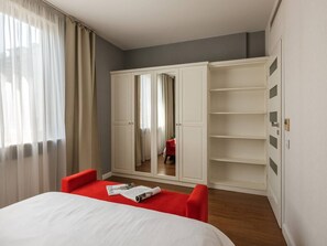 A hotel room with a bed, red chairs, a wardrobe, and a mirror.