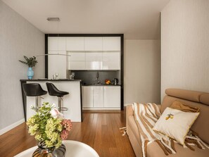 Un apartamento moderno con cocina, dos taburetes negros, un sofá con cojines a rayas y un jarrón con flores en el suelo.