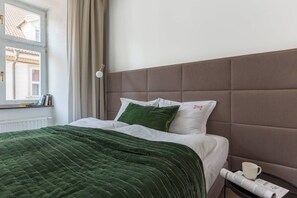 Ein ordentlich bezogenes Bett mit einer grünen Steude, einem braunen Polsterkopf und einem Nachttisch mit Tasse und Zeitung.