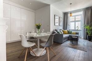 A modern living room with a dining area, grey sofa, and a large window with curtains.
