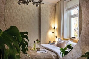 A bedroom with a large plant, a bedside table with a lamp, and a window with curtains.