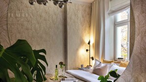 A bedroom with a large plant, a bedside table with a lamp, and a window with curtains.