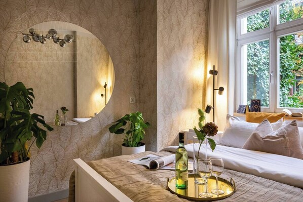 A hotel room with a large mirror, a bed with white linens, a bedside table with a tray holding a bottle and glasses, and a window with a view of greenery.