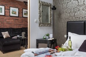 A modern room with a brick wall, a black sofa, a bed with white bedding, a small table with a lamp, and a mirror.