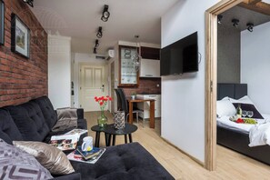 A modern living room with a sofa, a small table with books and a mug, a kitchen area with a dining table, and a bedroom with a bed and a TV.