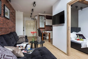 A modern living room with a sofa, a small table with books and a mug, a kitchen area with a dining table, and a bedroom with a bed and a TV.