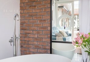A bathroom with a brick wall, a modern shower, and a view of a living area with a sofa and flowers. - Glamour studio with free-standing bathtub (Kraków)