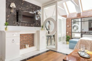 A modern living room with a fireplace, a TV mounted on a marble wall, a circular mirror, and a dining area with a table set for two. - Glamour studio with free-standing bathtub (Kraków)