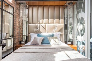 A modern bedroom with a tufted headboard, a large bed with patterned bedding, and a glass partition with a decorative mirror.