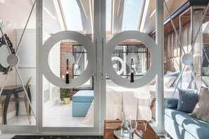 A modern interior with a glass door leading to an outdoor seating area, a round mirror, and a wooden table with glasses.
