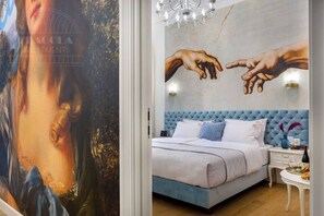Une chambre à coucher avec un grand tableau, une tête de lit recouverte d’un tissu bleu, un couvre-lit blanc et une table de chevet sur laquelle se trouvent un vase et une bouteille de vin.