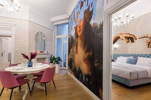 A modern interior with a dining area, a large painting, and a bedroom with a tufted headboard.