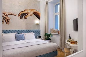 A bedroom with a large bed, a wall mural, a window with curtains, a bedside table with a bottle and glasses, and a plant.