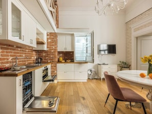 A compact kitchen with a brick wall, white cabinetry, a dining table with chairs, and a flat-screen TV.