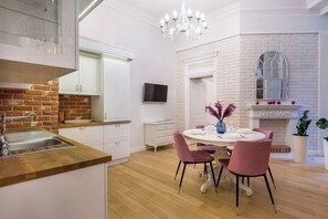 Une cuisine moderne avec des meubles blancs, un mur d’accent en briques, un coin repas avec des chaises roses et une cheminée.