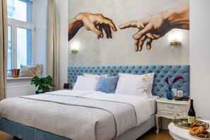 A bedroom with a large bed, a wall mural, a window, and a bedside table with a vase and books.