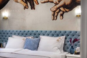 Une chambre à coucher avec une tête de lit capitonnée, du linge de lit blanc, des coussins à motifs, une fresque murale et une table de chevet ornée d’un vase et d’un livre.