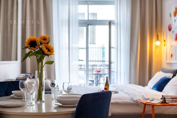 A hotel room with a dining table set for two, a vase with sunflowers, a bed with white and blue bedding, and a balcony view.