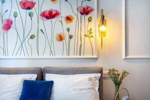A bedroom with a bed, pillows, a wall-mounted light, and a floral painting.